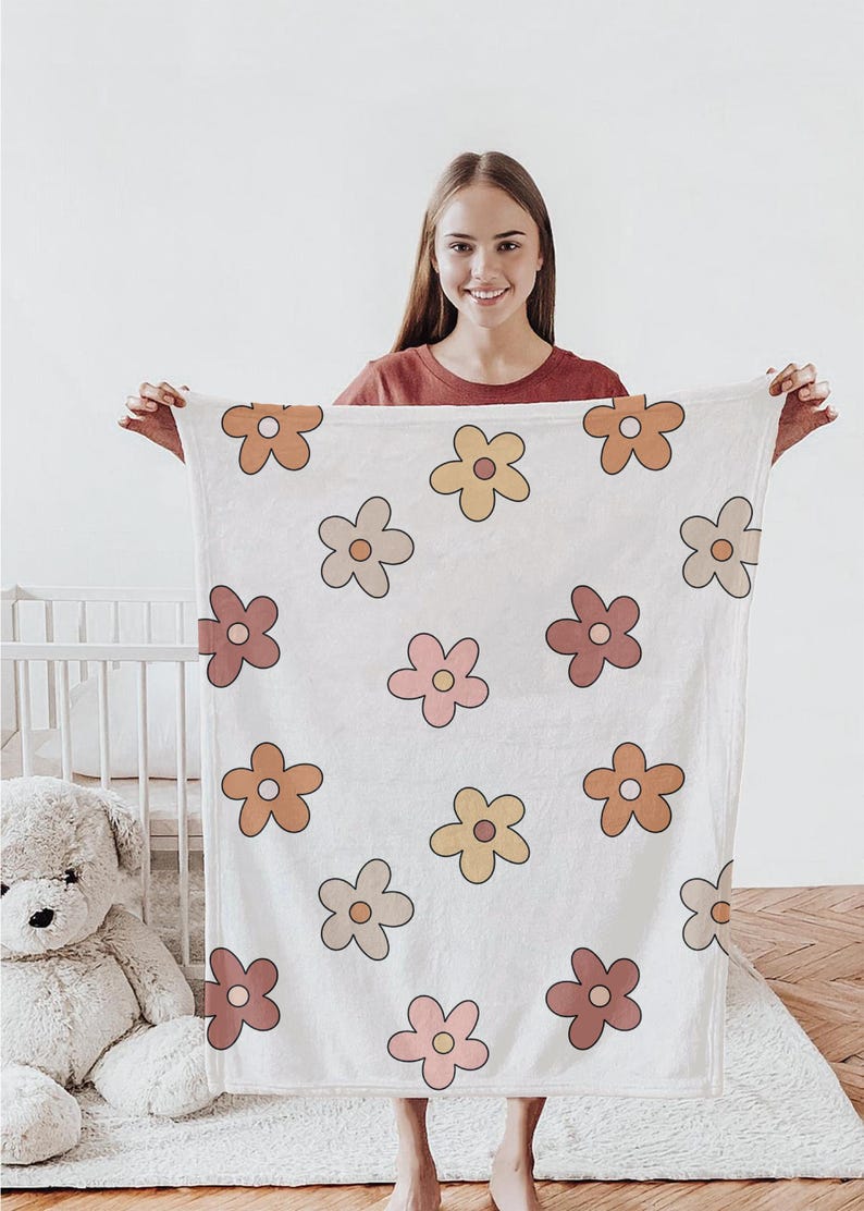 K&ouml;nnte beinhalten: Eine wei&szlig;e Babydecke mit einem Blumenmuster mit orangefarbenen, gelben und rosa Blumen. Die Decke ist weich und flauschig, perfekt zum Kuscheln.