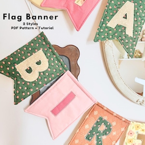 May include: A fabric flag banner featuring individual pennants in pink, green, and orange with floral patterns and appliqued letters. The banner displays the text Flag Banner 2 Styles PDF Pattern + Tutorial. Each pennant has a swallowtail bottom.