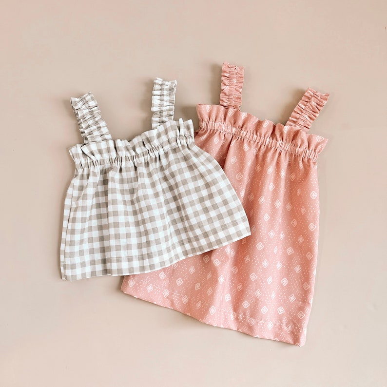 Puede incluir: Dos blusas sin mangas con tirantes con volantes. Una blusa es de cuadros marrones y blancos, la otra es rosa con un patr&oacute;n geom&eacute;trico blanco.
