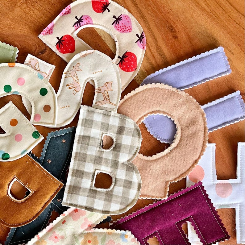 Pode incluir: Uma cole&ccedil;&atilde;o de letras de tecido coloridas, incluindo um "B" com um padr&atilde;o xadrez bege e branco, um "D" em bege e um "R" com desenhos de morangos. Outras letras apresentam bolinhas, estampas florais e uma cor castanho claro.