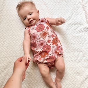 Op de afbeelding: Een baby die een roze en oranje bloemenprint sleeveless onesie draagt. De onesie heeft een gerimpelde rok en een ronde halslijn.