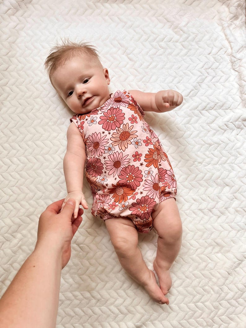 Op de afbeelding: Een baby die een roze en oranje bloemenprint sleeveless onesie draagt. De onesie heeft een gerimpelde rok en een ronde halslijn.