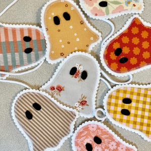 May include: A garland featuring seven fabric ghost shapes in various patterns and colours, including floral, plaid, and striped prints. Each ghost has two black felt oval eyes and is finished with a white scalloped edge, all connected by a white cord.