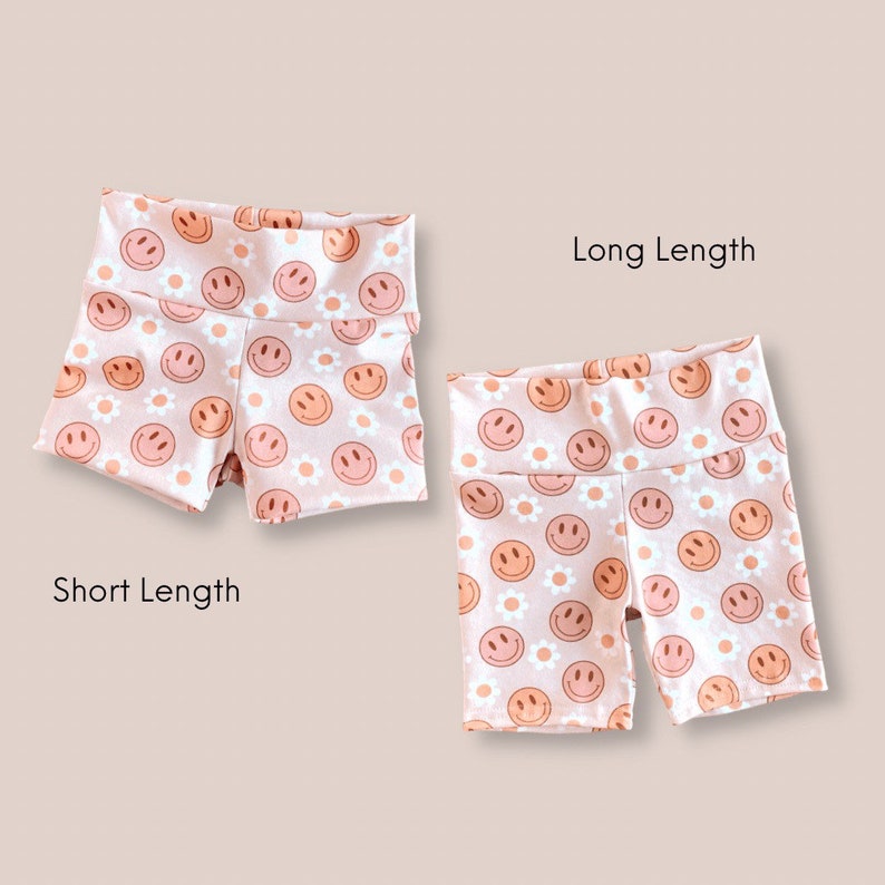 May include: Two pairs of pink shorts with a white smiley face and daisy print. The shorts are shown in two lengths, short and long.