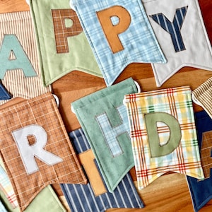 May include: A fabric banner featuring individual pennants with various patterns and colors, including plaid, stripes, and solid tones. Each pennant displays a stitched letter, collectively spelling out the phrase HAPPY BIRTHDAY in mixed fonts.
