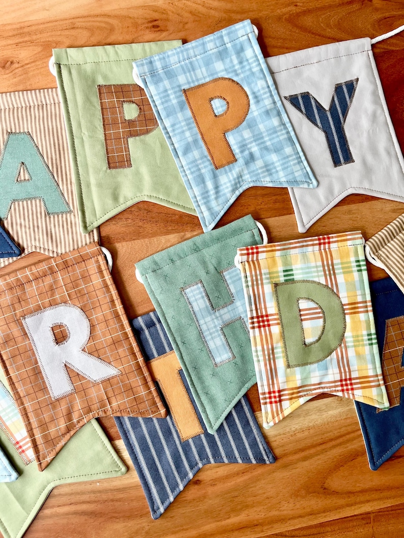 May include: A fabric banner featuring individual pennants with various patterns and colors, including plaid, stripes, and solid tones. Each pennant displays a stitched letter, collectively spelling out the phrase HAPPY BIRTHDAY in mixed fonts.