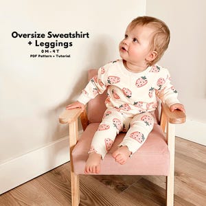 May include: A child wearing a cream-colored strawberry-patterned sweatshirt and leggings set. The outfit features pink strawberries with green leaves. The text "Oversize Sweatshirt + Leggings" and "0M-4T" is visible in the image.