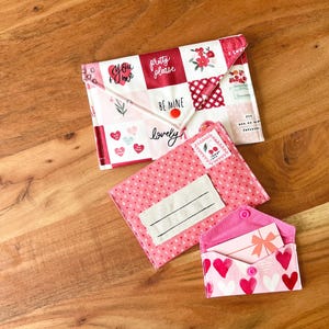 Op de afbeelding: Drie stoffen enveloppen in verschillende maten en patronen worden getoond op een houten oppervlak. De grootste envelop heeft de tekst "You + Me", "pretty please" en "BE MINE". De kleinste envelop is versierd met rode harten.