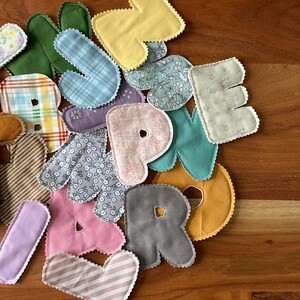 May include: A collection of colorful fabric letters in various patterns and colors, including yellow, pink, blue plaid, and floral prints. Each letter has a scalloped edge and is arranged on a wooden surface.