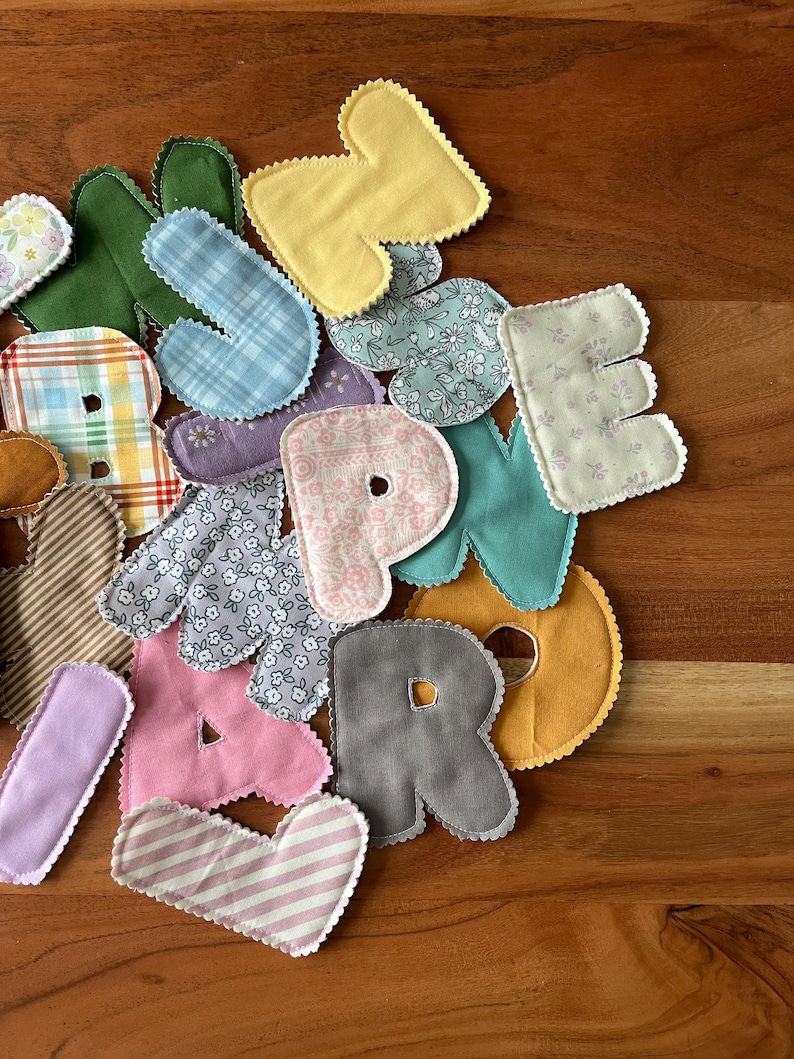 May include: A collection of colorful fabric letters in various patterns and colors, including yellow, pink, blue plaid, and floral prints. Each letter has a scalloped edge and is arranged on a wooden surface.