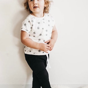 May include: A young child wearing a white short-sleeved t-shirt with a black dandelion print and black pants.
