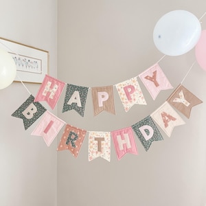 May include: A fabric banner spelling out HAPPY BIRTHDAY in two rows. The pennants are made of various solid and floral patterned fabrics in shades of pink, sage green, and tan. Each letter is appliqued onto the center of the individual pennant flags.