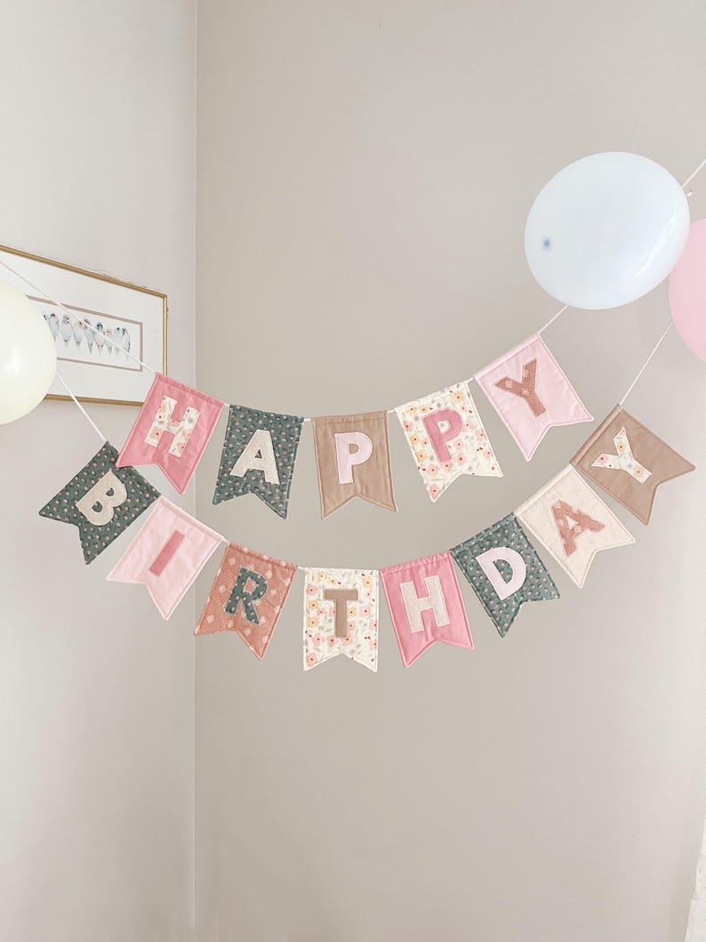 May include: A fabric banner spelling out HAPPY BIRTHDAY in two rows. The pennants are made of various solid and floral patterned fabrics in shades of pink, sage green, and tan. Each letter is appliqued onto the center of the individual pennant flags.