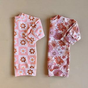 May include: Two long-sleeved shirts with a pink background and different patterns. One shirt has a checkered pattern with brown smiley faces. The other shirt has a floral pattern with brown and pink flowers.