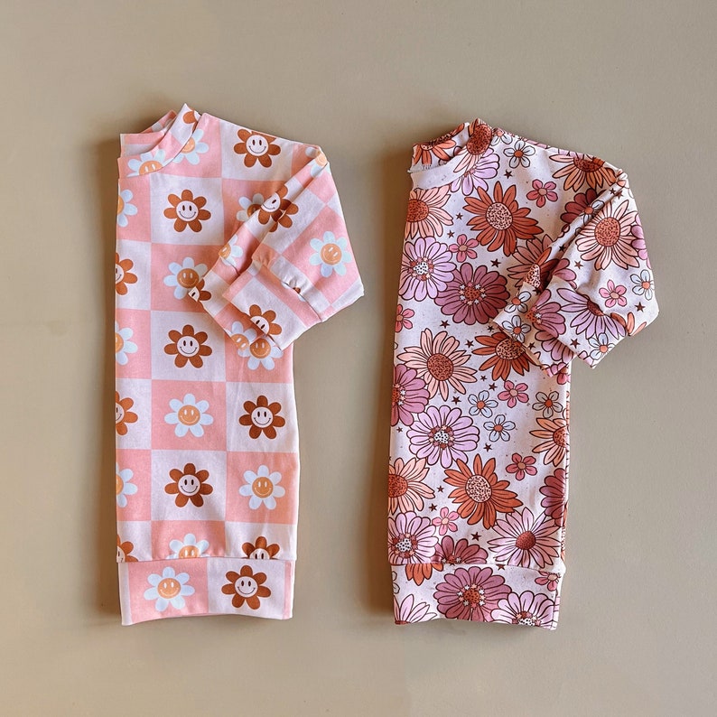 May include: Two long-sleeved shirts with a pink background and different patterns. One shirt has a checkered pattern with brown smiley faces. The other shirt has a floral pattern with brown and pink flowers.