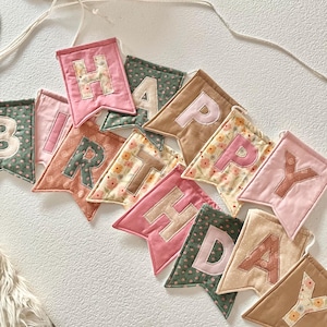 May include: A fabric banner spelling HAPPY BIRTHDAY with individual pennants in various colours including pink, green, tan, and cream. Each pennant features a letter in a contrasting colour, with some fabrics displaying floral patterns and others solid.