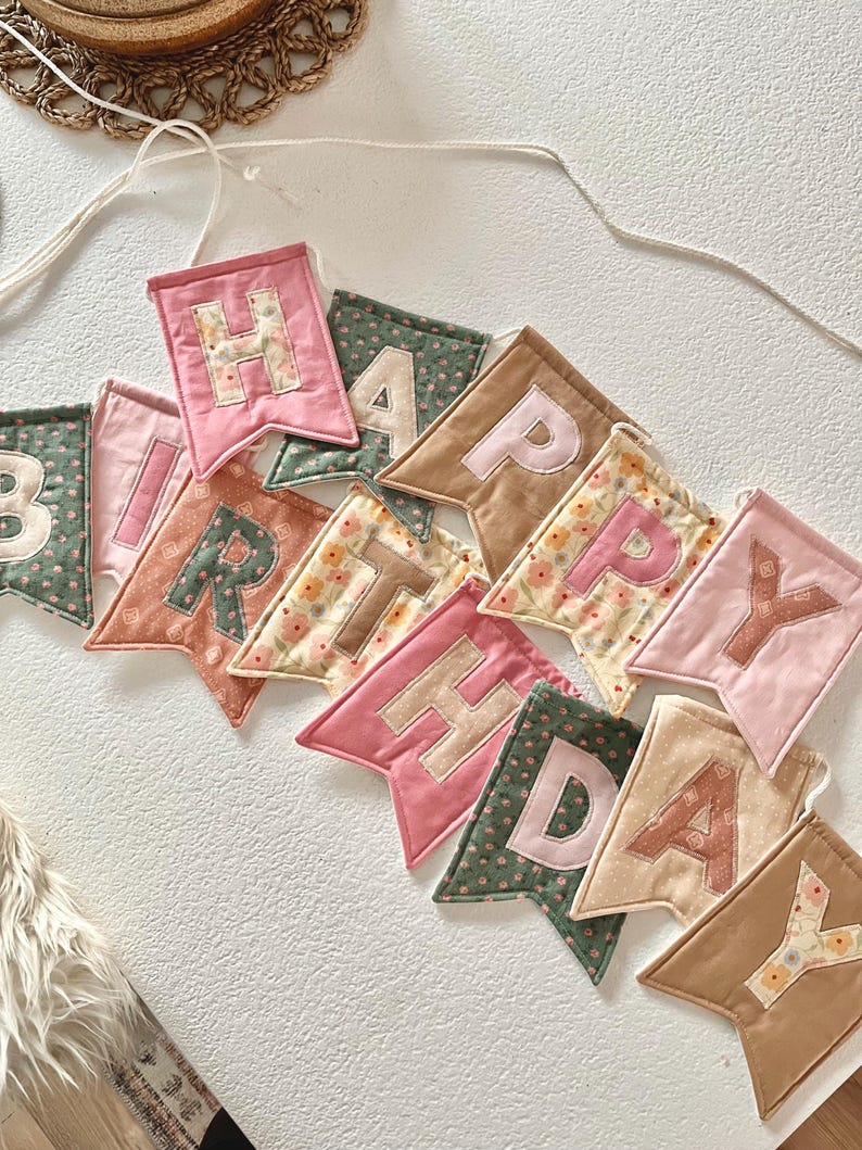 May include: A fabric banner spelling HAPPY BIRTHDAY with individual pennants in various colours including pink, green, tan, and cream. Each pennant features a letter in a contrasting colour, with some fabrics displaying floral patterns and others solid.