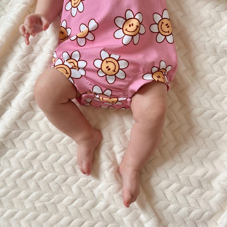 Op de afbeelding: Een roze baby-romper met een wit madeliefjespatroon. Elke madeliefje heeft een geel smiley-gezicht in het midden.