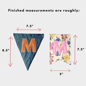 May include: Two fabric pennants with the text 'Finished measurements are roughly:'. The blue triangular pennant with a gold star pattern and orange letter 'M' measures 7.5 inches wide by 8.5 inches high. The floral rectangular pennant with a pink letter 'M' measures 5 inches wide by 7.5 inches high.