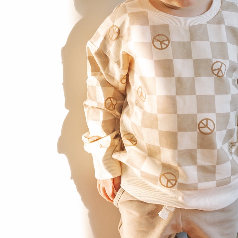 Puede incluir: Una sudadera de color beige y blanco a cuadros con s&iacute;mbolos de paz marrones impresos en ella. La sudadera tiene mangas largas y cuello redondo.