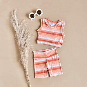 May include: A striped tank top and matching shorts in shades of pink and orange. The outfit is laid out on a light brown surface with a pair of white sunglasses.