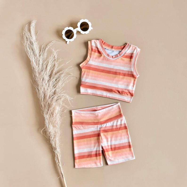 May include: A striped tank top and matching shorts in shades of pink and orange. The outfit is laid out on a light brown surface with a pair of white sunglasses.