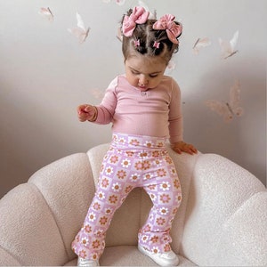 May include: A young child wearing a pink long-sleeved shirt and pink and white floral bell-bottom pants. The child is sitting on a white fluffy chair.