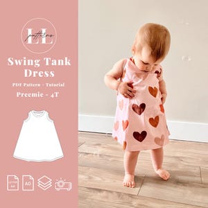 Può includere: Un vestito a trapezio rosa chiaro per bambini piccoli, decorato con cuori marroni e arancioni di varie dimensioni. L'abito è indossato da un bambino piccolo in piedi su un pavimento di legno. L'immagine include anche il testo "Swing Tank Dress" e "Preemie - 4T".