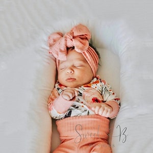 May include: A baby wearing a pink floral print onesie with long sleeves and matching pink ribbed leggings. The baby is wearing a large pink bow headband. The baby is sleeping on a white pillow.
