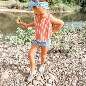 May include: A young child wearing a red and white striped tank top and blue floral shorts stands on a rocky surface. She wears a blue floral headband and light green sandals. She holds a yellow sunflower.