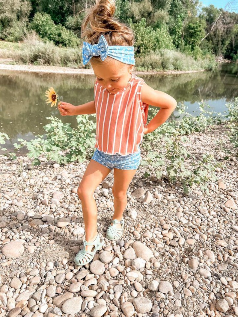 May include: A young child wearing a red and white striped tank top and blue floral shorts stands on a rocky surface. She wears a blue floral headband and light green sandals. She holds a yellow sunflower.