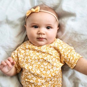 K&ouml;nnte beinhalten: Ein Baby tr&auml;gt einen gelben und wei&szlig;en Blumen-Overall mit einem gelben Schleifen-Haarband.