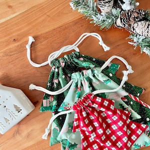 May include: A collection of Christmas-themed drawstring bags in various patterns. The bags feature festive designs including gift boxes, holly, and plaid. Each bag has a white drawstring closure. The bags are arranged on a wooden surface, with a decorative candle holder and pine boughs in the background.