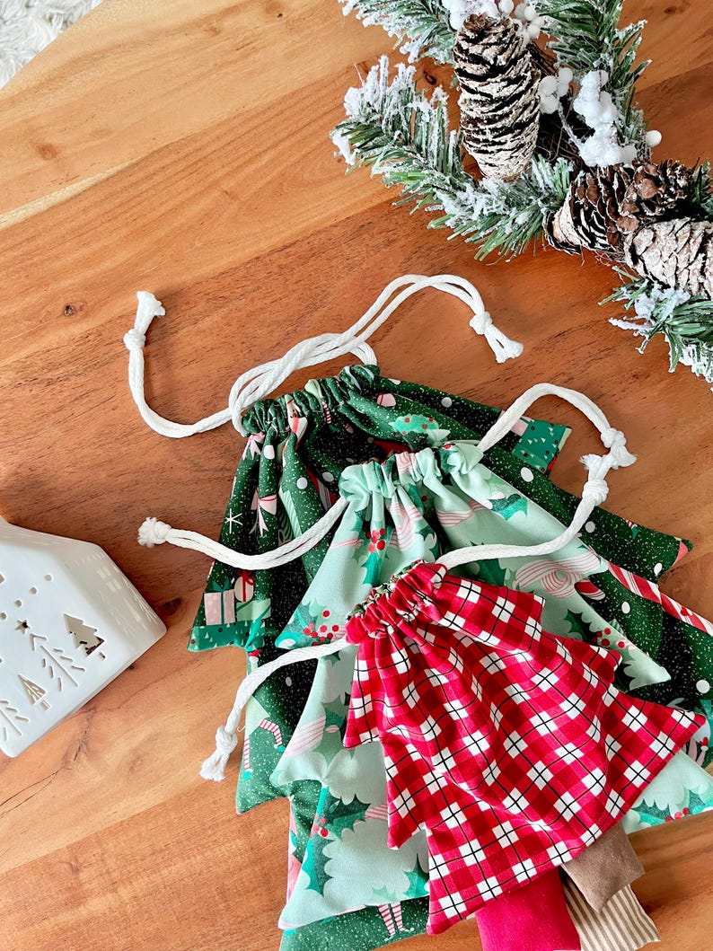 May include: A collection of Christmas-themed drawstring bags in various patterns. The bags feature festive designs including gift boxes, holly, and plaid. Each bag has a white drawstring closure. The bags are arranged on a wooden surface, with a decorative candle holder and pine boughs in the background.