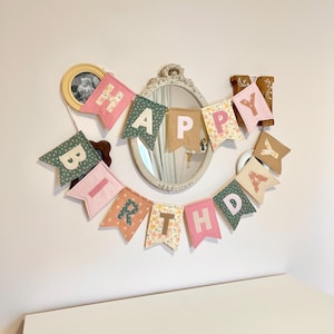 May include: A fabric banner spelling out HAPPY BIRTHDAY with individual pennant flags in various colours and patterns including pink, green, tan, and floral prints. Each flag features a letter in a contrasting colour, hung in a curved arrangement.