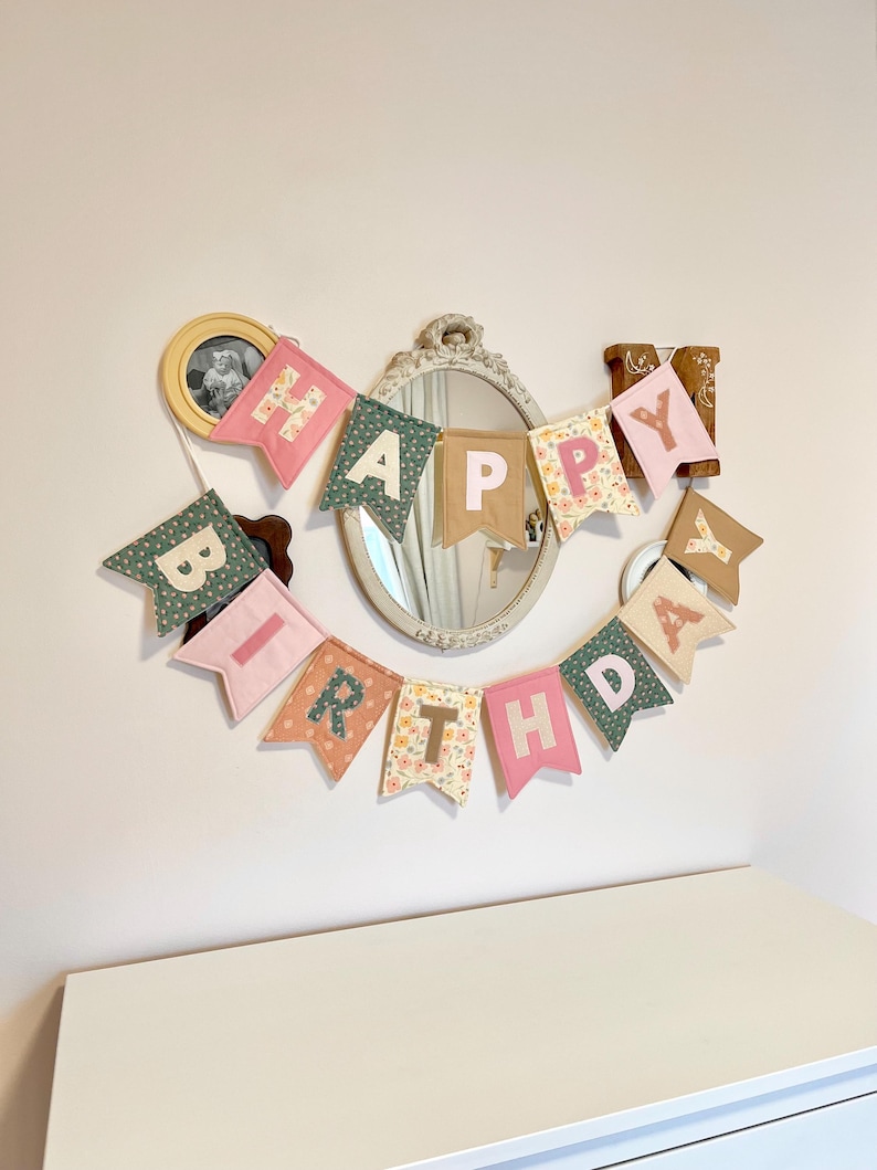 May include: A fabric banner spelling out HAPPY BIRTHDAY with individual pennant flags in various colours and patterns including pink, green, tan, and floral prints. Each flag features a letter in a contrasting colour, hung in a curved arrangement.