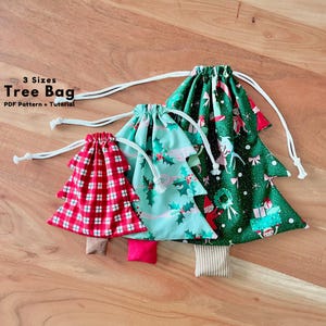 May include: Three tree-shaped drawstring bags in three sizes. The smallest bag features a red and white plaid pattern. The medium bag has a light green background with a holly leaf print. The largest bag is dark green with a holiday elf print.