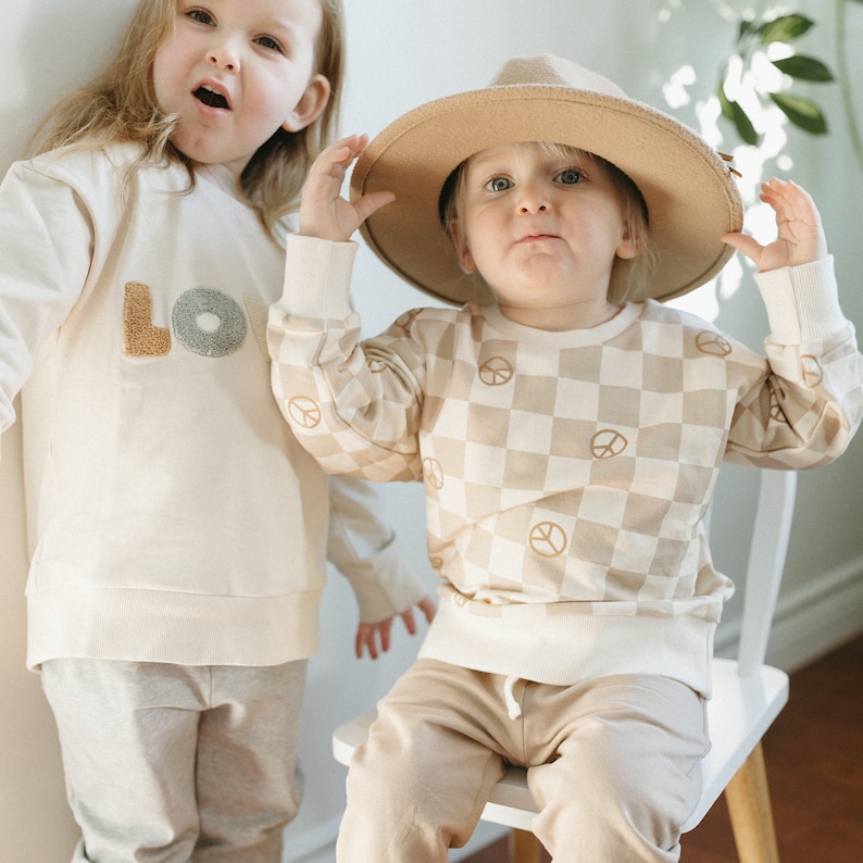 Puede incluir: Dos ni&ntilde;os con sudaderas de color crema. El ni&ntilde;o de la izquierda lleva una sudadera con la palabra "LOVE" bordada en la parte delantera. El ni&ntilde;o de la derecha lleva un sombrero marr&oacute;n y una sudadera a cuadros con s&iacute;mbolos de paz.