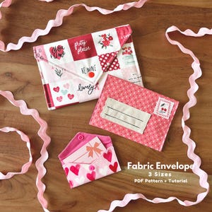 May include: Three fabric envelopes in different sizes and patterns, including hearts, floral, and text. The envelopes are pink and white, with a red button closure on the largest one. The text "Fabric Envelope" and "3 Sizes" is visible.