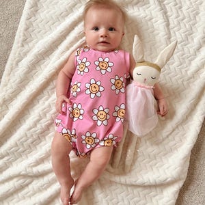 Op de afbeelding: Een baby draagt een roze, mouwloze romper met een witte madeliefjesprint. De romper heeft gele smiley-gezichten in het midden van elke madeliefje. De baby houdt een roze en witte knuffelkonijn vast.