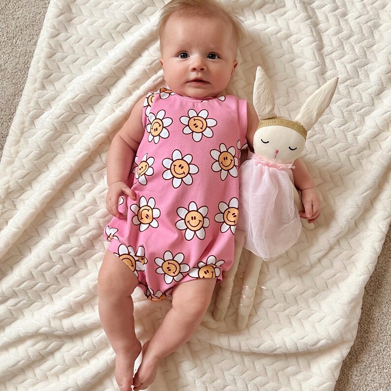 Op de afbeelding: Een baby draagt een roze, mouwloze romper met een witte madeliefjesprint. De romper heeft gele smiley-gezichten in het midden van elke madeliefje. De baby houdt een roze en witte knuffelkonijn vast.