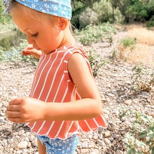 May include: A young child wearing a light blue floral headband, a coral-colored top with white vertical stripes, and blue floral shorts. The outfit is designed for warm weather and outdoor activities.