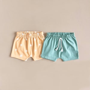 May include: Two pairs of toddler shorts. One pair is a light yellow with a white sun pattern. The other pair is a solid light blue with a white drawstring.