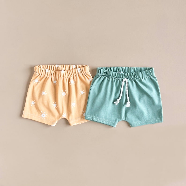 May include: Two pairs of toddler shorts. One pair is a light yellow with a white sun pattern. The other pair is a solid light blue with a white drawstring.