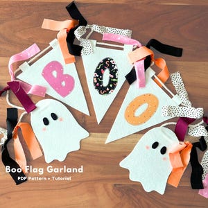 Può includere: Una ghirlanda decorativa con tre bandierine triangolari in feltro bianco con le lettere B, O e O, e due sagome di fantasmi in feltro bianco. Le lettere sono rosa, nere con motivi di caramelle e arancioni. La ghirlanda è decorata con nastri in tessuto nero, arancione e rosa e strisce di tessuto a pois. Il testo "Boo Flag Garland PDF Pattern + Tutorial" è visibile.