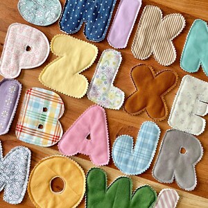 May include: A collection of colorful, fabric-covered letters with scalloped edges. The letters are in various patterns and colors, including floral, polka dot, and striped designs. The letters spell out a word, perfect for a nursery or craft project.