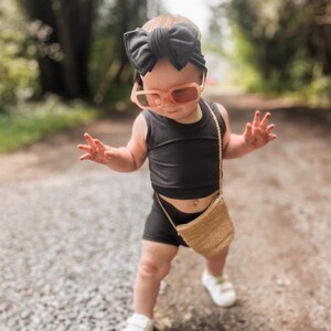 May include: A toddler wearing a black sleeveless top, black shorts, and a black bow headband. The toddler is also wearing pink sunglasses and a small brown woven bag.