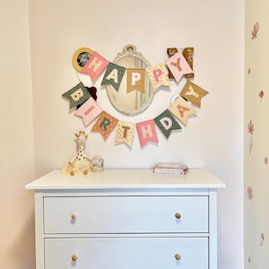 May include: A fabric birthday banner featuring individual pennants in various colours and floral patterns, spelling out HAPPY BIRTHDAY in white letters. The banner is hung on a wall above a white three-drawer dresser, positioned around an oval mirror.