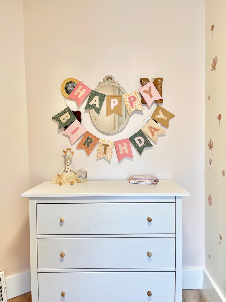 May include: A fabric birthday banner featuring individual pennants in various colours and floral patterns, spelling out HAPPY BIRTHDAY in white letters. The banner is hung on a wall above a white three-drawer dresser, positioned around an oval mirror.
