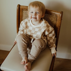 Puede incluir: Un ni&ntilde;o peque&ntilde;o lleva un su&eacute;ter a cuadros marr&oacute;n y blanco con s&iacute;mbolos de paz impresos. El ni&ntilde;o est&aacute; sentado en una silla de madera con un asiento de ca&ntilde;a tejido. El ni&ntilde;o lleva pantalones marrones.